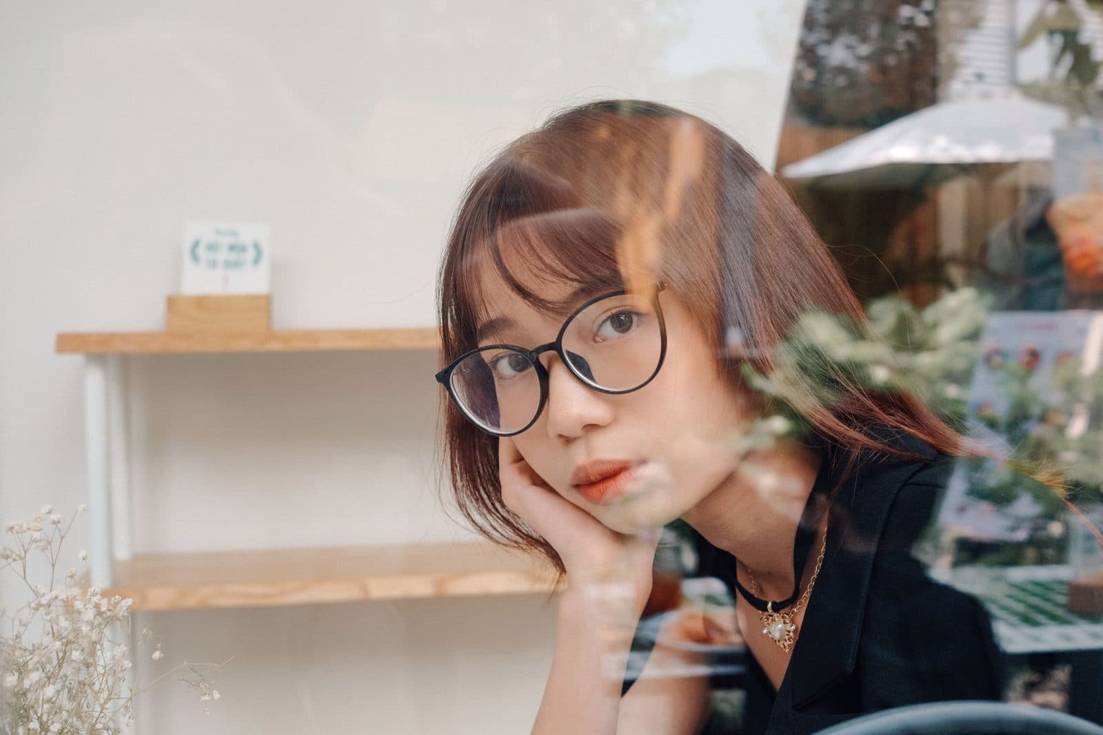 Cô gái đeo gọng kính tròn chống cằm sau ô kính quán cà phê