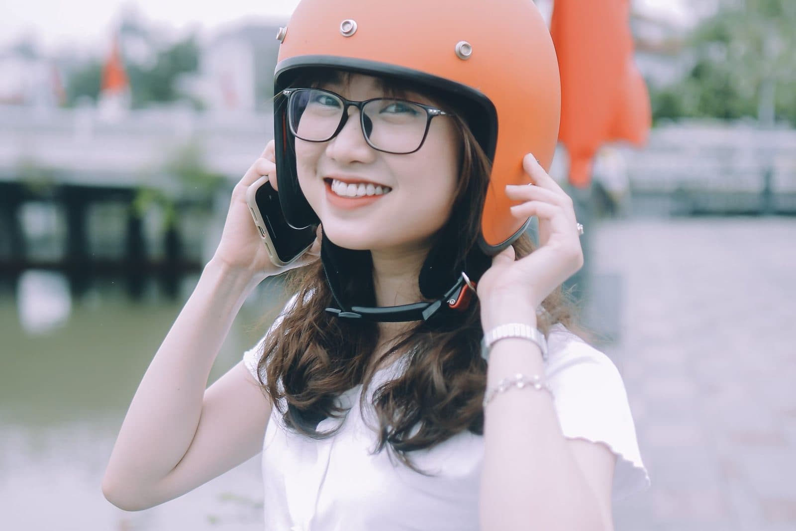 Cô gái đội mũ bảo hiểm cam, đeo gọng kính đen, cười tươi bên bờ sông